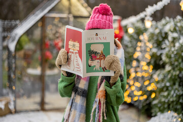 Young woman reads some magazine on New Year's theme at snowy backyard decorated for a winter...