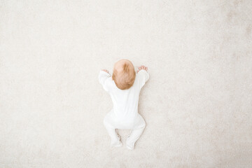 Baby in white bodysuit crawling on knee and arms on light beige home carpet. 5 to 6 months old infant development. Top down view. - obrazy, fototapety, plakaty