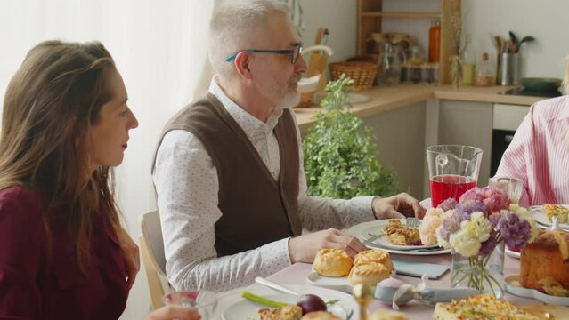Panning shot of family chatting and clinking drinks in toast while little girl playing on digital tablet during Easter dinner at home