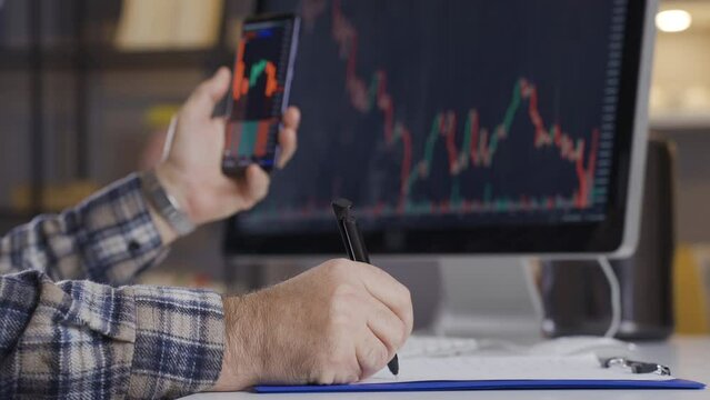 Professional trader is following the stock market and taking notes from phone and computer.
The trader looks and analyzes the charts on his computer and phone. Stocks and cryptocurrency.
