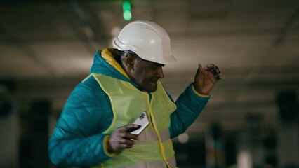 Active energetic mature cheerful man professional builder engineer foreman in protective helmet and uniform rhythmic dancing funny moving to dynamic music on mobile phone celebrates winning great deal