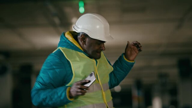 Active energetic mature cheerful man professional builder engineer foreman in protective helmet and uniform rhythmic dancing funny moving to dynamic music on mobile phone celebrates winning great deal