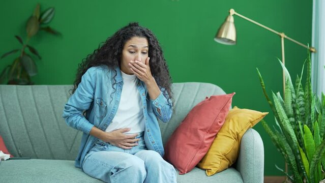 Sick young dark haired curly woman suffering from stomach ache feeling bad nausea sitting on sofa at home Unhealthy female lady holding belly with poisoning vomiting reflex pregnancy syndrome indoors 