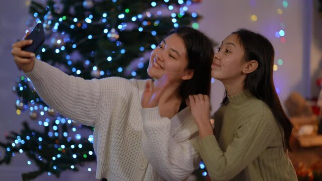 Young happy Asian women grimacing gesturing air kiss waving taking selfie on Christmas eve indoors. Charming positive female friends photographing with smartphone app smiling on New Year's eve