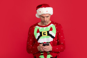 Santa chatting with phone. Portrait of middle aged man in sweater isolated over red background. Concept of holidays, happiness, emotions and Christmas celebration.