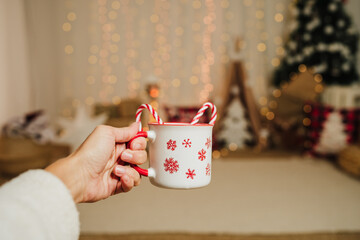 woman hand holding christmas mug with hot drink and candy indoor over christmas decoration