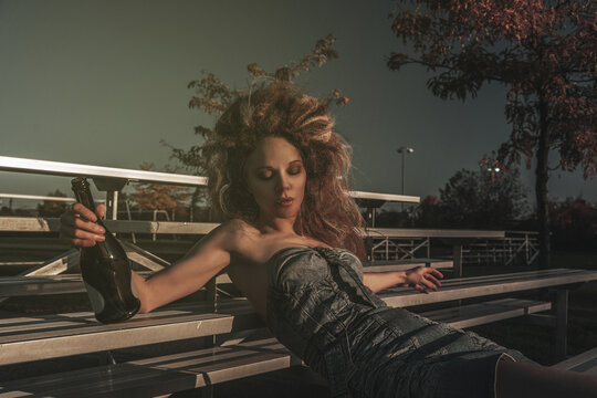 Vintage portrait of a tipsy beautiful young woman wearing an old school denim dress and long teased hair, celebrating alone with champagne in the bleachers of a soccer field