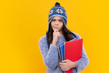 Thinking pensive clever teenager girl. School children in winter hat and sweater with books on isolated yellow studio background. Children learning and education.