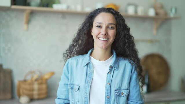 Smiling dark haired curly woman with crossed hands looking at camera while standing on the light domestic kitchen indoor Portrait of charming happy hispanic housewife lady posing at home alone 