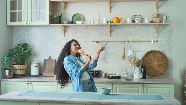 Happy energetic curly woman singing karaoke song in spoon and dancing on light domestic kitchen in slow motion indoor Young dark haired hispanic female relaxing and having fun alone at home lifestyle 