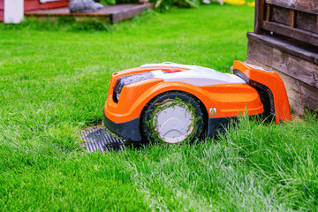 Automatic lawn robot mower under rain at charging stations on the grass, lawn. Close up side view with blurry background - obrazy, fototapety, plakaty