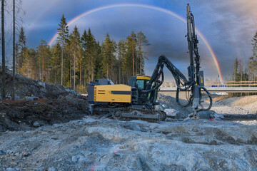 Yellow drilling machine under colorful rainbow in sky stands on granite rock and drills holes for...