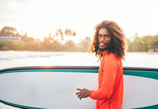 Portrait of black long-haired teen boy with surfboard ready for surfing with sunset backlight. He walking in Indian ocean waves. Extreme water sports and exotic countries concept, Udawalawe, Sri Lanka