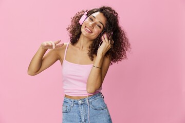 Happy woman wearing headphones with curly hair listening to music and singing along with her eyes closed in a pink T-shirt and jeans on a pink background DJ party, copy space