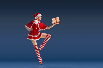 Dancing ballerina with a gift in her hands on pointe shoes in the costume of Santa Claus on a dark blue background.
