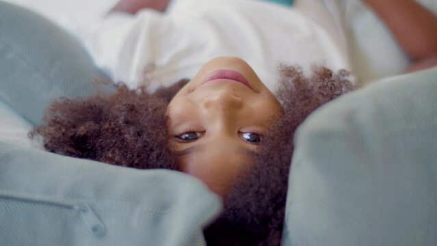 Face closeup of cute Black girl lying in bed looking at camera. Pretty child with Black curly hair enjoying leisure time, resting her head between pillows. Static shot. Childhood concept.