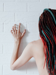 Naked girl with long dreadlocks in her hair standing near a white brick wall with her hand raised