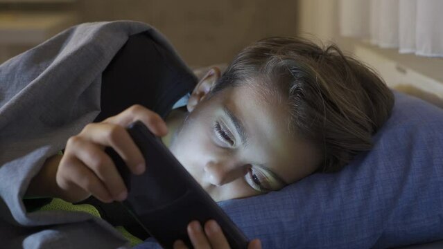Phone addicted boy plays with sleepless phone. 
The child, who can hardly open his eyes from lack of sleep, falls asleep while playing on the phone in his bed at night.
