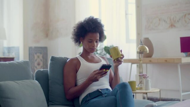 Young Black Woman Drinking Tea While Web Surfing On Her Mobile Phone, Sitting On Cozy Couch At Home. Pretty African American Lady Using Mobile App, Sipping Coffee. Modern Technology, Lifestyle Concept