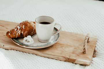 Wooden tray with cup of coffee, fresh croissant with white frosting and white flower on ceramic plate - romantic breakfast in bed
