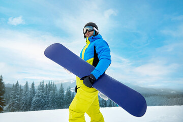 Portrait Of Man On The Background Of Mountains And Blue Sky, Holding Snowboard And Wearing Ski...