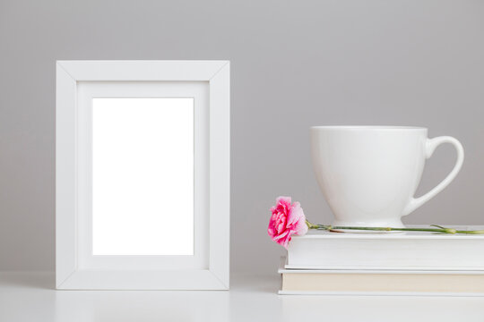 White picture frame mockup on desk with books, porcelain white mug and pink flower	- romantic still life composition