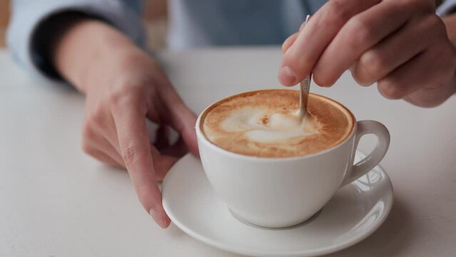 Close-up of a cappuccino with thick foam white cup of hot drink in the hands of a white woman stirs coffee in a cup with a metal spoon, a manager's lunch break is sitting in a cafe restaurant
