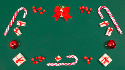 Christmas greeting card. Rectangular frame of candy canes, christmas balls, twigs with red berries and gifts boxes on viridian green background.