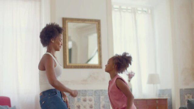 Happy Black mother and daughter dancing at home together. Attractive African American woman showing dance moves to cute little girl, standing opposite her. Slow motion, side view. Happy family concept
