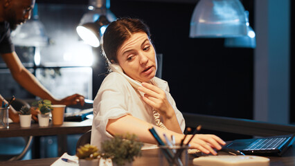 Female freelancer answering landline phone call in office, working late at night to create business...