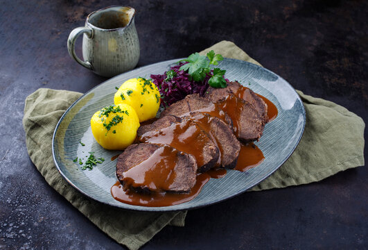 Traditionell geschmorter Sauerbraten vom Rind mit Blaukraut und Kartoffelkl&ouml;&szlig;en in w&uuml;rziger Dunkelbierso&szlig;e serviert als close-up auf einem Nordic design Teller