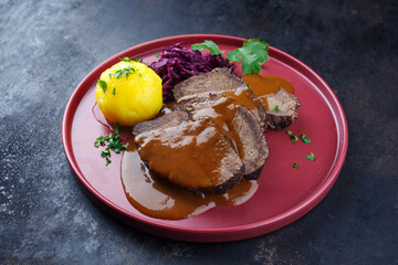 Traditionell geschmorter Sauerbraten vom Rind mit Blaukraut und Kartoffelklößen in würziger Dunkelbiersoße serviert als close-up auf einem Nordic design Teller