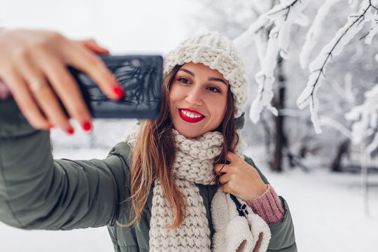 Portrait of young woman taking selfie on smartphone in snowy winter park. Girl wearing warm clothes. Close up