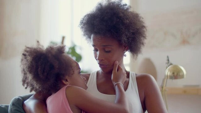 Young Black woman embracing her daughter, touching her face and smiling while comforting child with kind words. Little girl stroking moms cheek. Happy family, motherhood, mother and daughter concept.