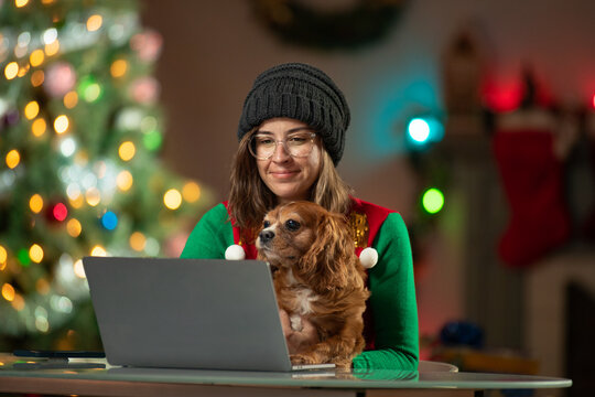 Young woman holding her Cavalier King Charles Spaniel in front of a computer. Christmas Tree in the background of her living room.