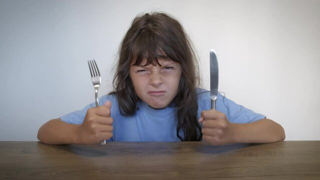 Angry child with knife and fork indoor. An angry little girl ask for food with knife and fock by the empty table in the room. A concept of food crisis and economical inflation in the world.