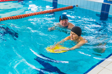 Swimming lessons concept. Caucasian female pensioner in swimming gear learning how to swim with the use of swimming kickboard. supported by her senior adult male friend. . High quality photo - Powered by Adobe