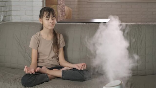 Girl meditation and aromatherapy indoor. A teenage girl practicing aromatherapy and meditation on the sofa in the room with working purifier.