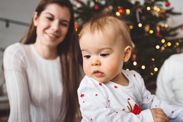 Cute baby girl in Christmas outfit, with blurred Christmas tree and her mom in the background