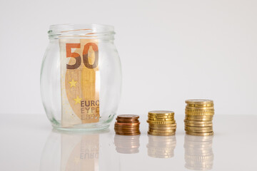 Stacked euro coins with on the side a glass jar with fifty euro banknote inside on a white background. Euro money saving concept. Copy space.