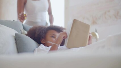 Little girl spending time reading in bed when her mum coming up, smiling and kissing childs forehead. Black woman talking to her daughter lying in bed, stroking her cheek gently. Parenthood concept.