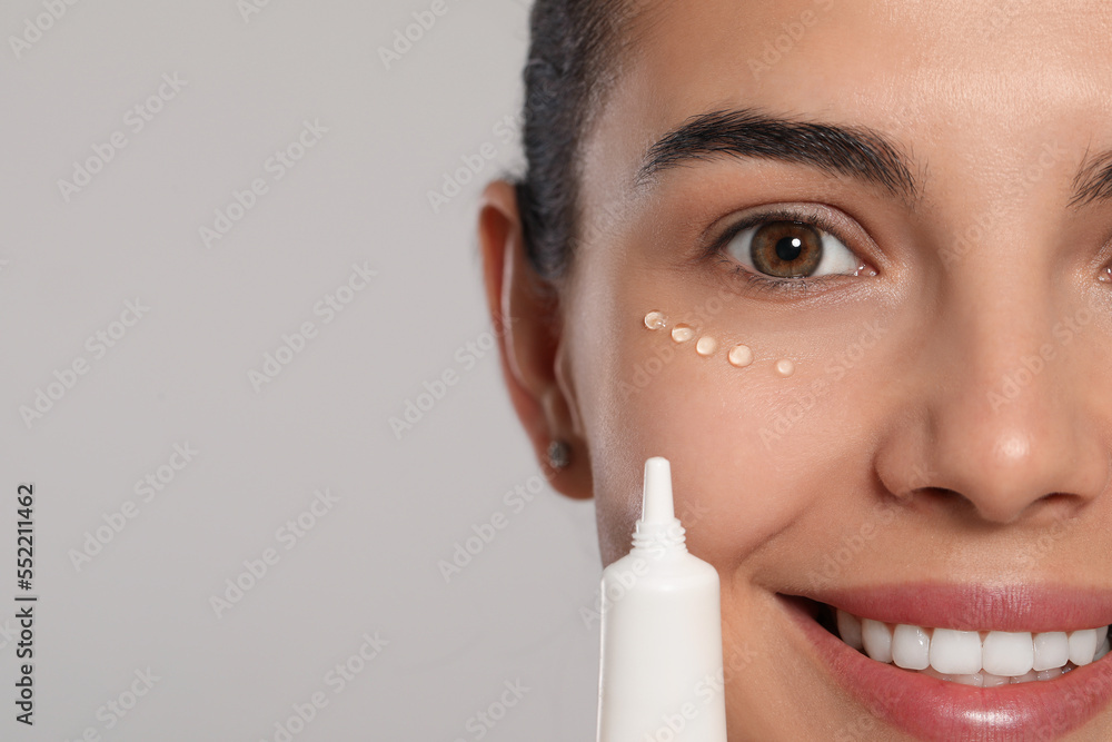 Wall mural Beautiful young woman applying gel on skin under eye against light grey background, closeup. Space for text