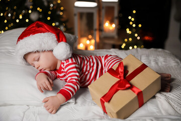 Baby in Christmas pajamas and Santa hat sleeping near gift box on bed indoors