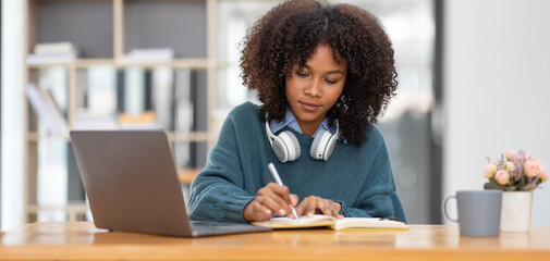 Cute african american female student wearing headphones with afro dreadlocks, studying remotely from home, using a laptop, taking notes on notepad during online lesson, e-learning concept, 