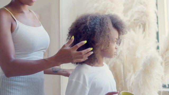 Cute Black girl standing drinking tea while mom making her hair, stroking daughters head gently. Thoughtful child touching soft toy and looking in window. Static shot. Childhood, parenthood concept.