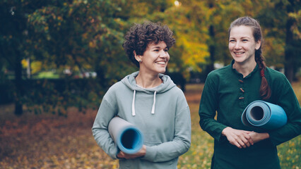 Beautiful girl is walking in park with yoga instructor holding mats talking gesturing and laughing....