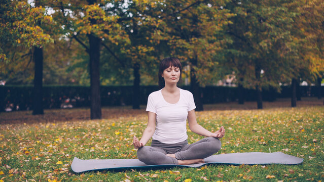 Attractive Young Lady Is Sitting In Lotus Position On Yoga Mat In Park Holding Hands In Mudra On Knees And Breathing Relaxing After Practice. Meditation, People And Nature Concept.