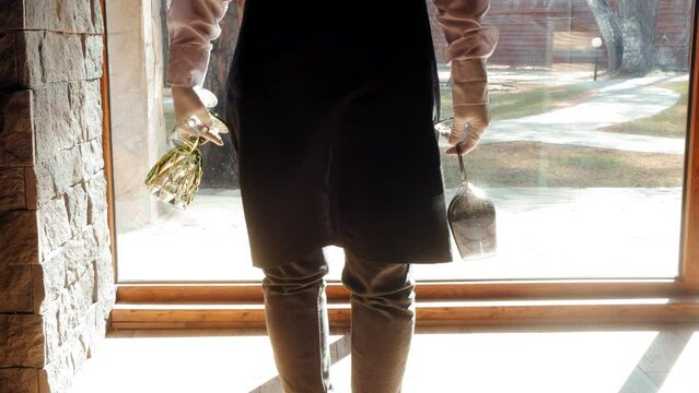 Unrecognizable waiter in uniform carrying wineglasses shining on sun with sunflare light leak, hands close-up. He is standing near big window in restaurant. Work of the service staff in modern cafe.