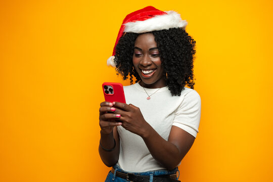 Attractive african lady in Santa hat using her cellphone on yellow background