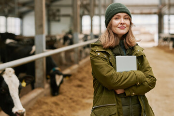 Portrait of young female agronomist with digital tablet looking at camera standing in barn with cows